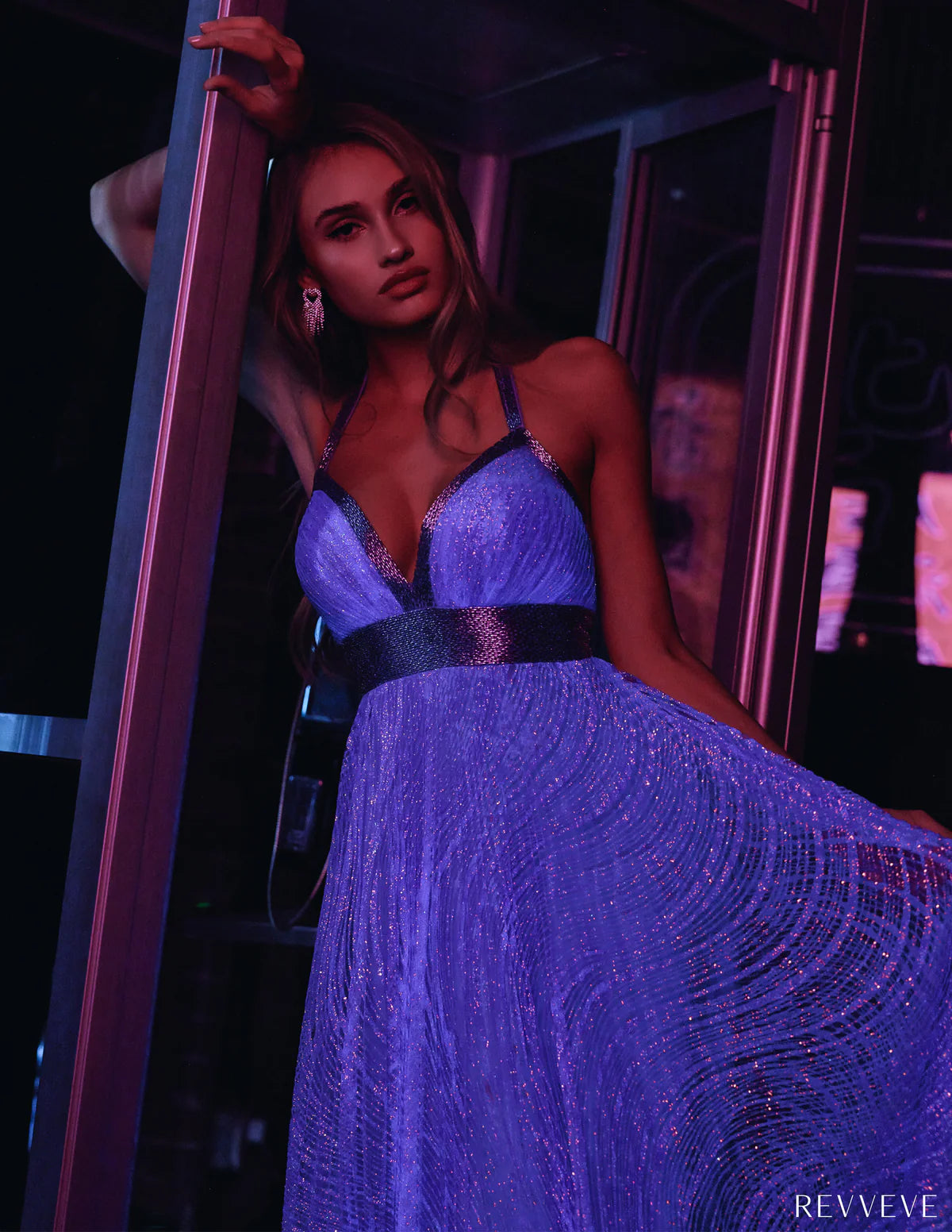 Stylish woman in a shimmering powder blue gown with glittering details, leaning against a doorway in a dimly lit room illuminated by vibrant purple and pink lighting.